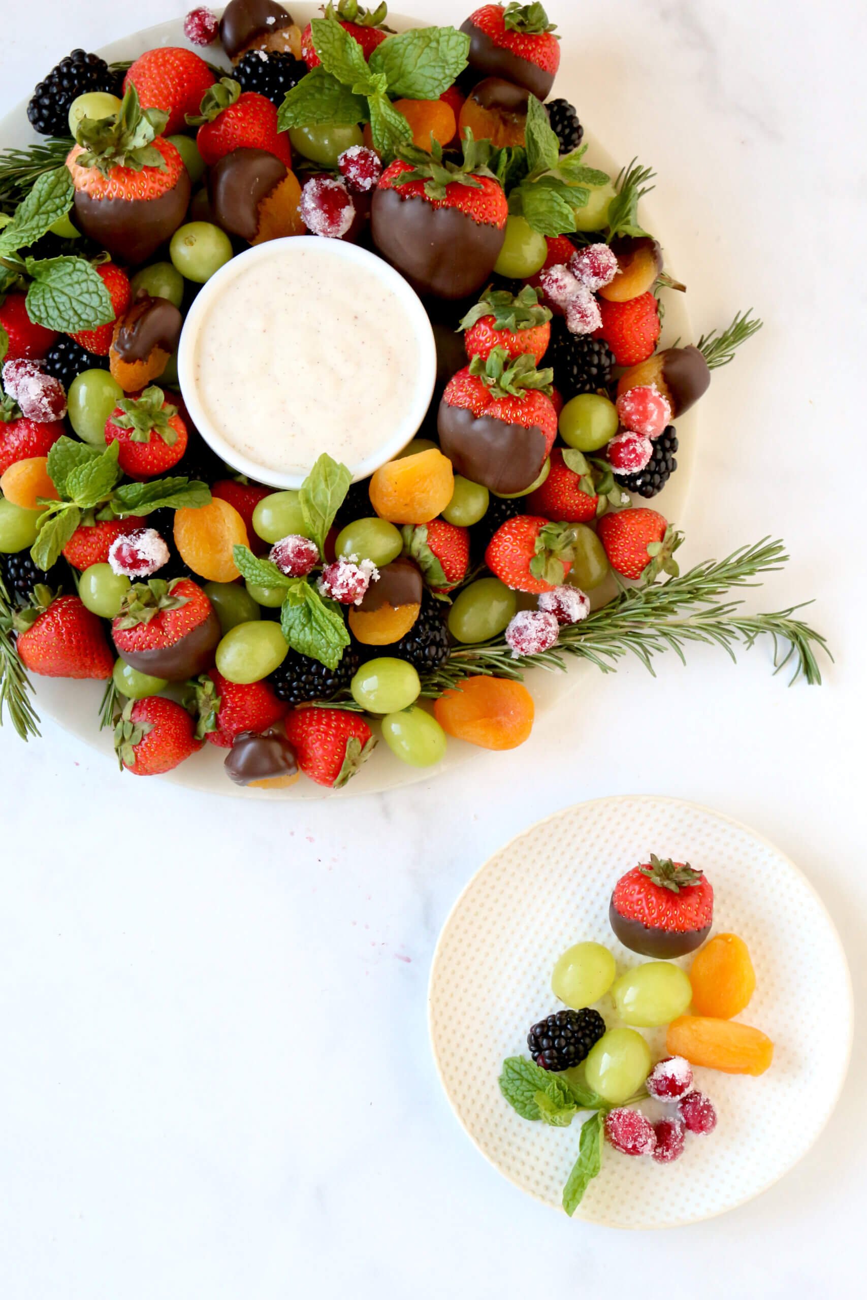 A round white platter topped with fresh strawberries, blackberries, green grapes, orange dried apricots and green fresh mint.