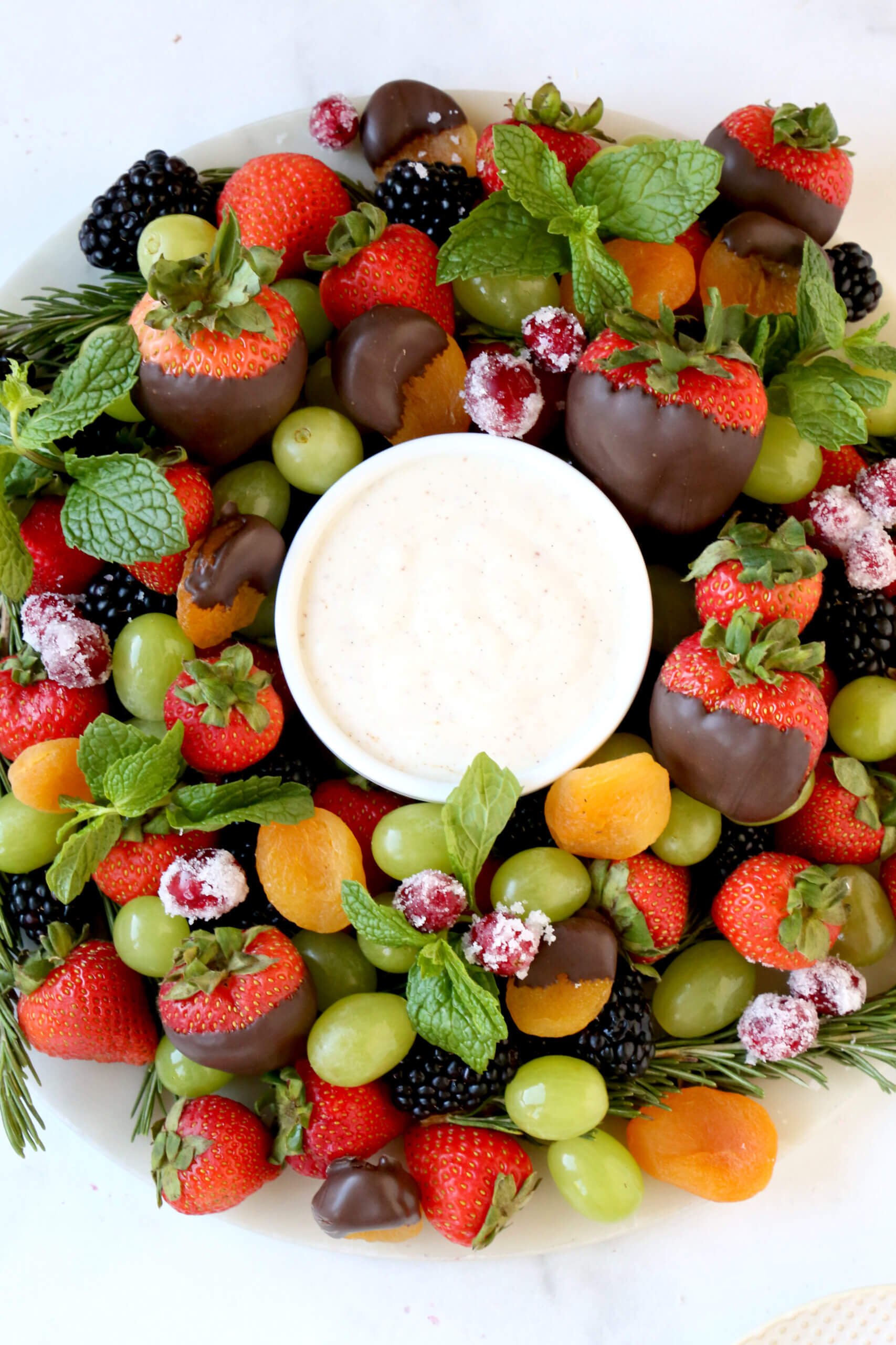 A round white platter topped with fresh strawberries, blackberries, green grapes, orange dried apricots and green fresh mint.