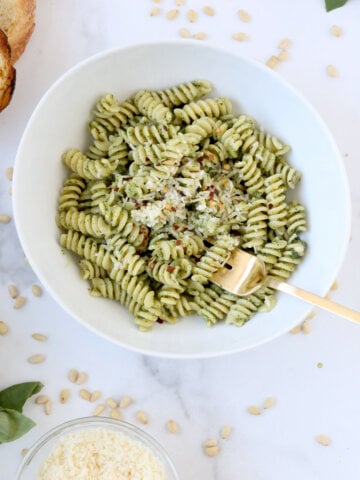 A white bowl with green pasta and a gold fork surrounded by white cheese and toasted bread.