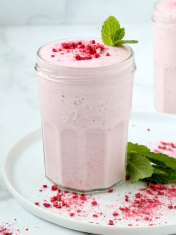 A clear glass filled with a pink creamy liquid, topped with a dark pink powder and a green mint leaf.