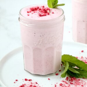 A clear glass filled with a pink creamy liquid, topped with a dark pink powder and a green mint leaf.