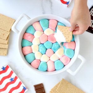 A white baking dish filled with red, white and blue marshmallows with a graham cracker dipped in.