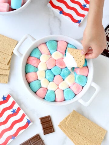 A white baking dish filled with red, white and blue marshmallows with a graham cracker dipped in.