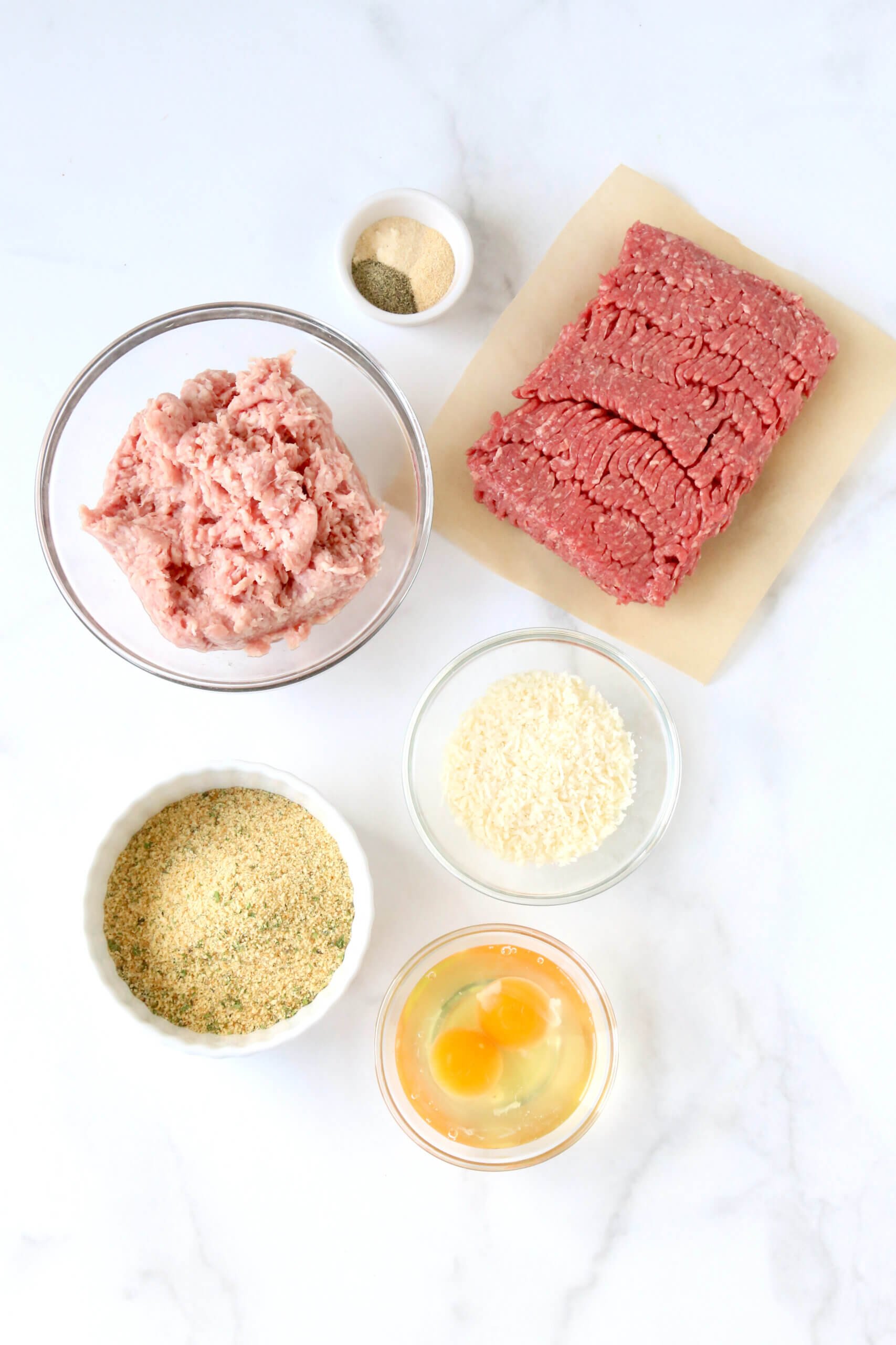 A square of red ground beef a lighter brown meat in a bowl, a bowls of tan bread crumbs next to a bowl of three eggs and white Parmesan cheese.  