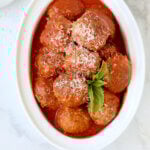 A white serving bowl filled with red tomato sauce and brown round meatballs with a green basil leaf on top.