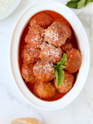 A white serving bowl filled with red tomato sauce and brown round meatballs with a green basil leaf on top.