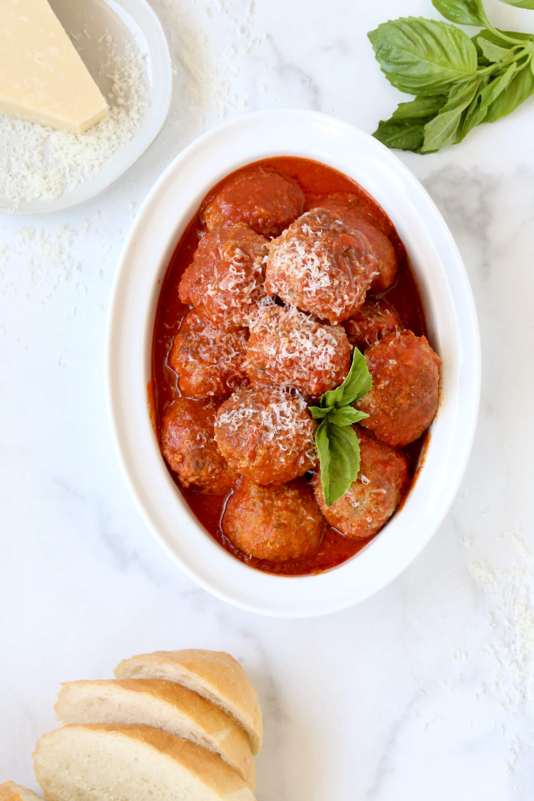A white serving bowl filled with red tomato sauce and brown round meatballs with a green basil leaf on top.