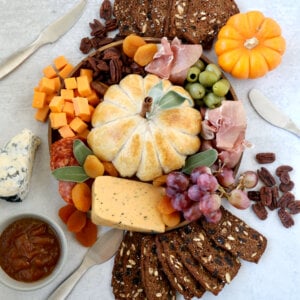 A brown round dish topped with a pumpkin looking baked brie, orange cheese cubes, prosciutto, salami, red grapes and crackers.