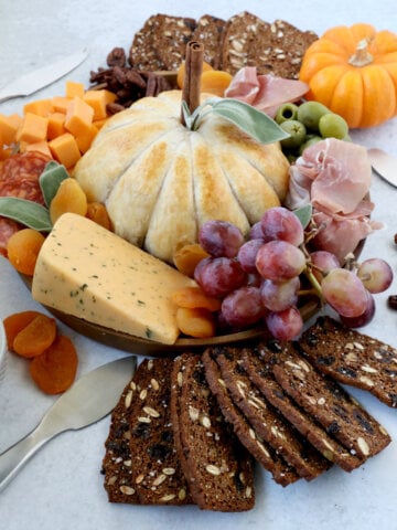 A brown round dish topped with a pumpkin looking baked brie, orange cheese cubes, prosciutto, salami, red grapes and crackers.