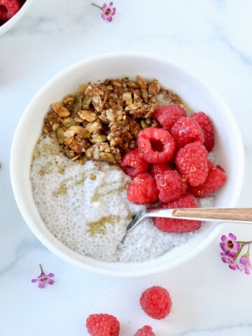 A white bowl filled with white pudding, fresh raspberries and light brown granola pieces. Next to a few raspberries and purples flowers.