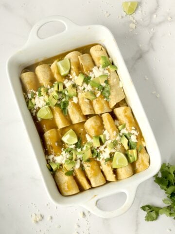 A white pyrex dish with light brown corn tortillas rolled up with light green sauce, green avocado diced and green cilantro on top.