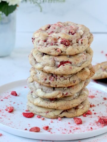 A stack of 6 cookies on a white plate with red crushed freeze dried strawberries sprinkled around it.