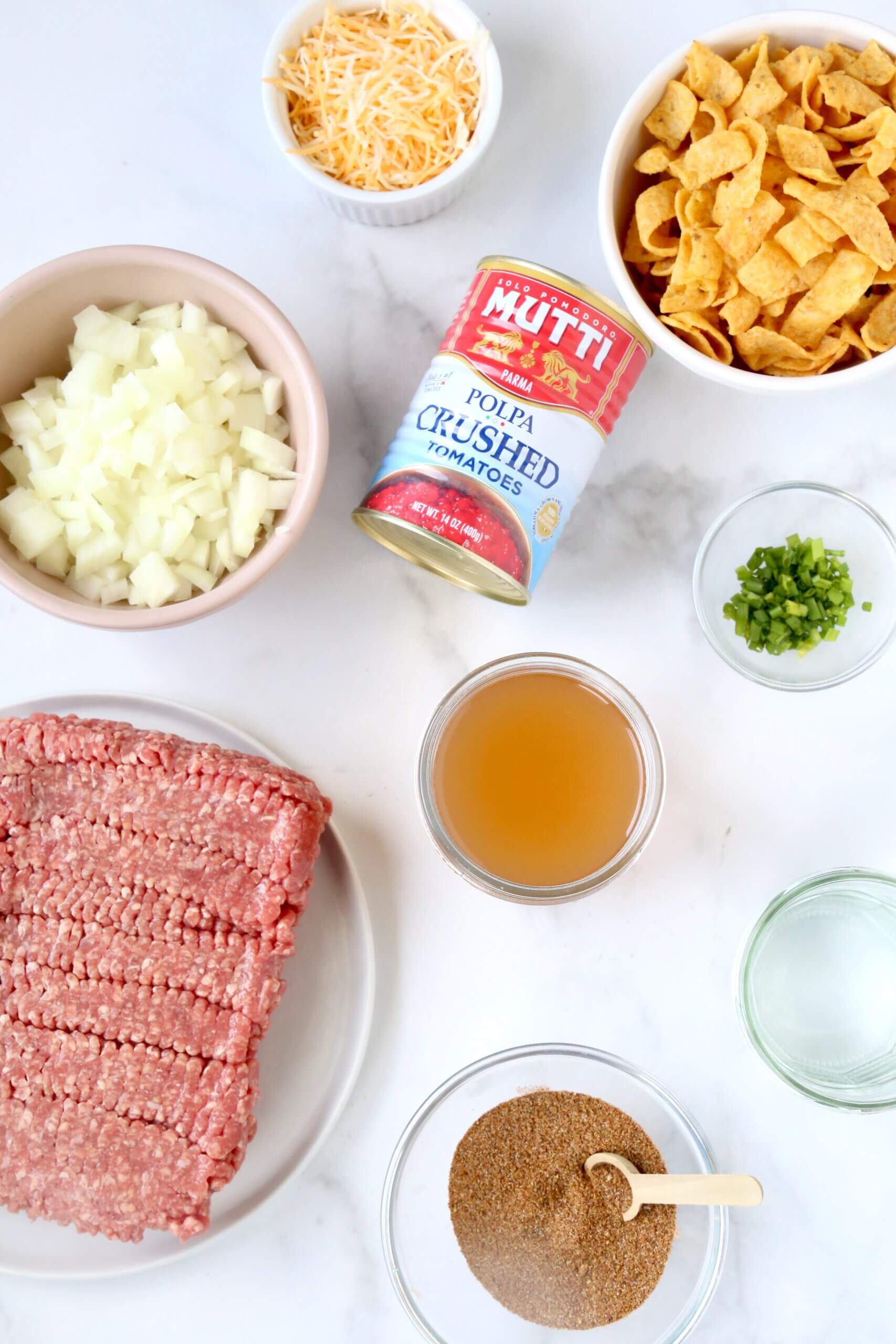 A photo of the ingredients included a square of red ground beef, a bowl of white chopped onions, a can of crushed tomatoes, a white bowl of yellow corn chips, a clear bowl of light brown beef broth and another clear bowl with green chopped chives.