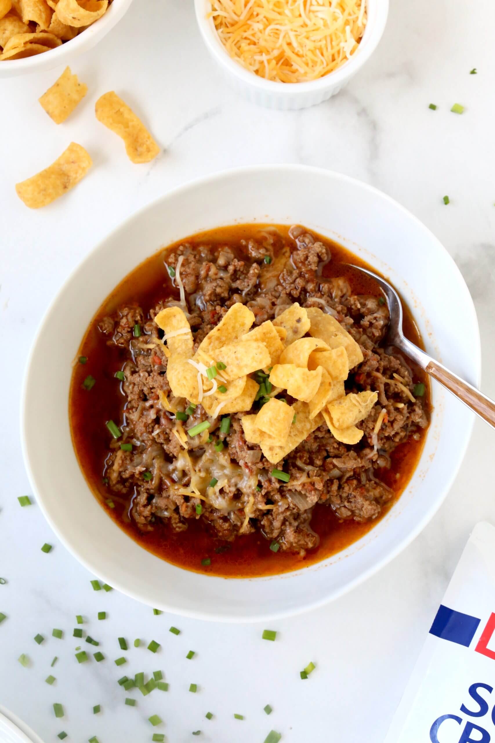 A white bowl with medium brown ground beef chili inside, topped with yellow corn chips and green chives.