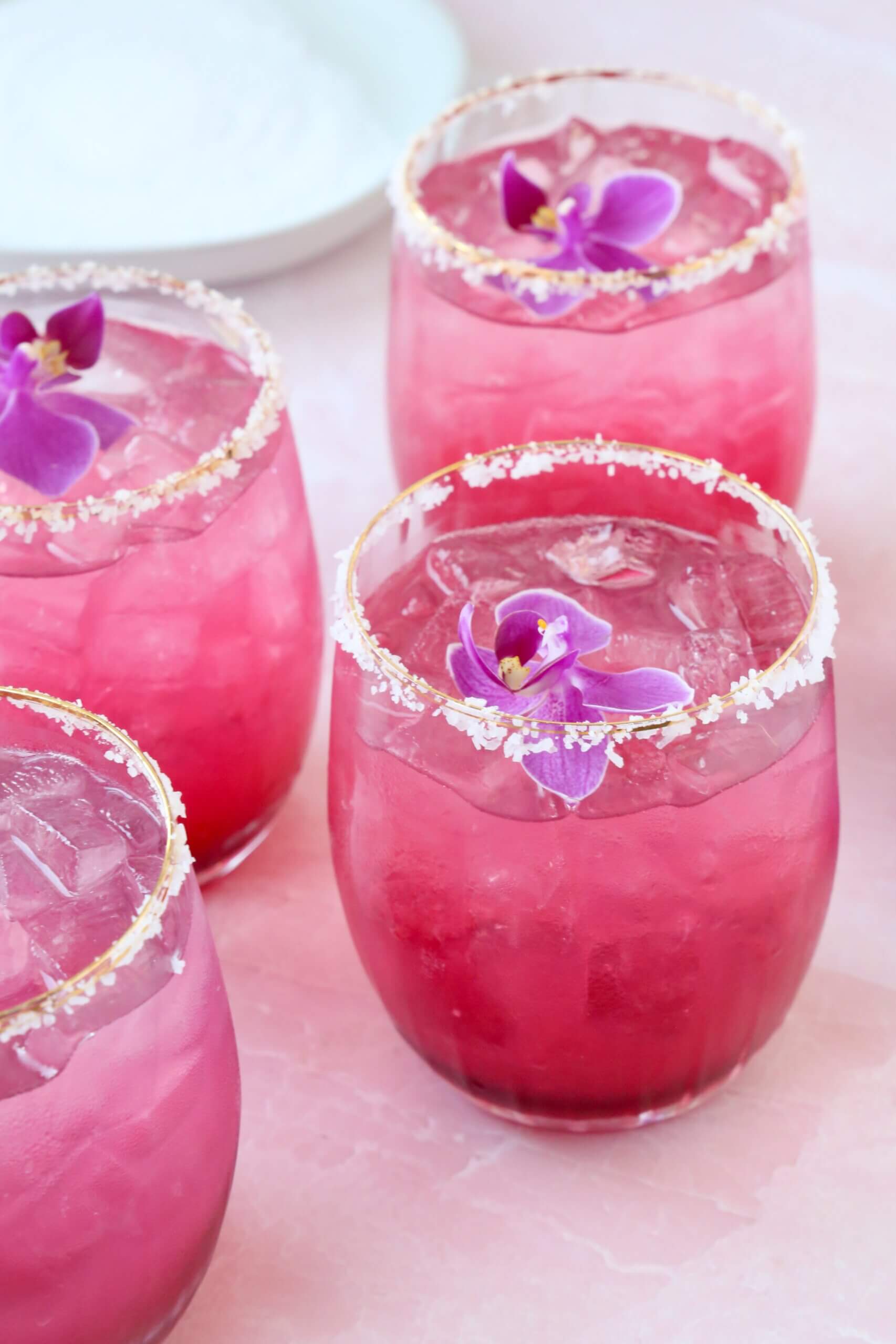 A clear glass with salt on the rim and a bright pink liquid and a purple orchid flower on top.