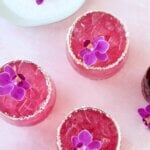 A clear glass with salt on the rim and a bright pink liquid and a purple orchid flower on top.