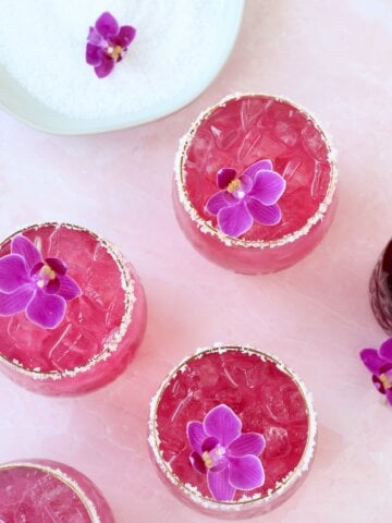 A clear glass with salt on the rim and a bright pink liquid and a purple orchid flower on top.