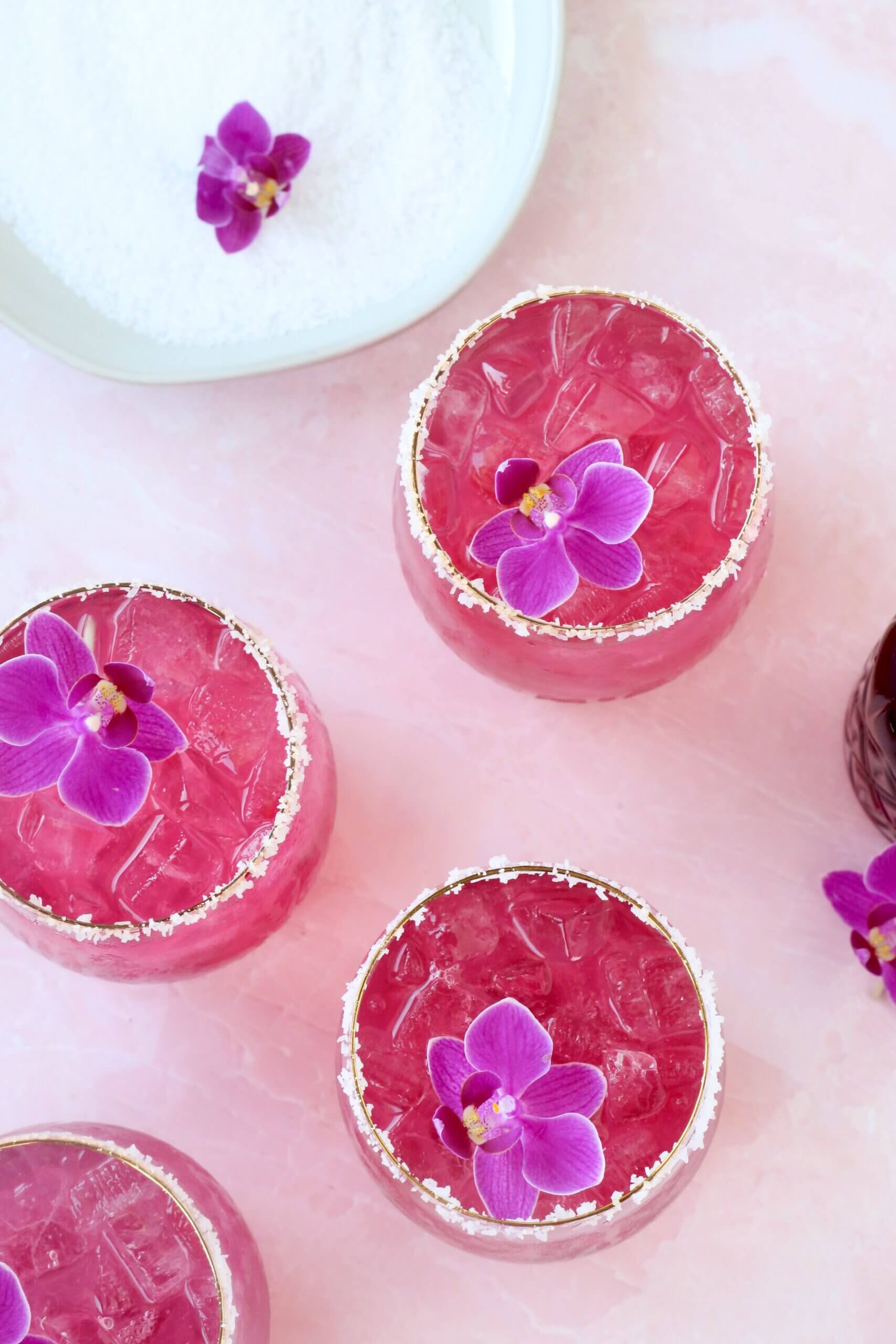 A clear glass with salt on the rim and a bright pink liquid and a purple orchid flower on top.