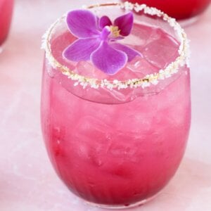 A clear glass with salt on the rim and a bright pink liquid and a purple orchid flower on top.