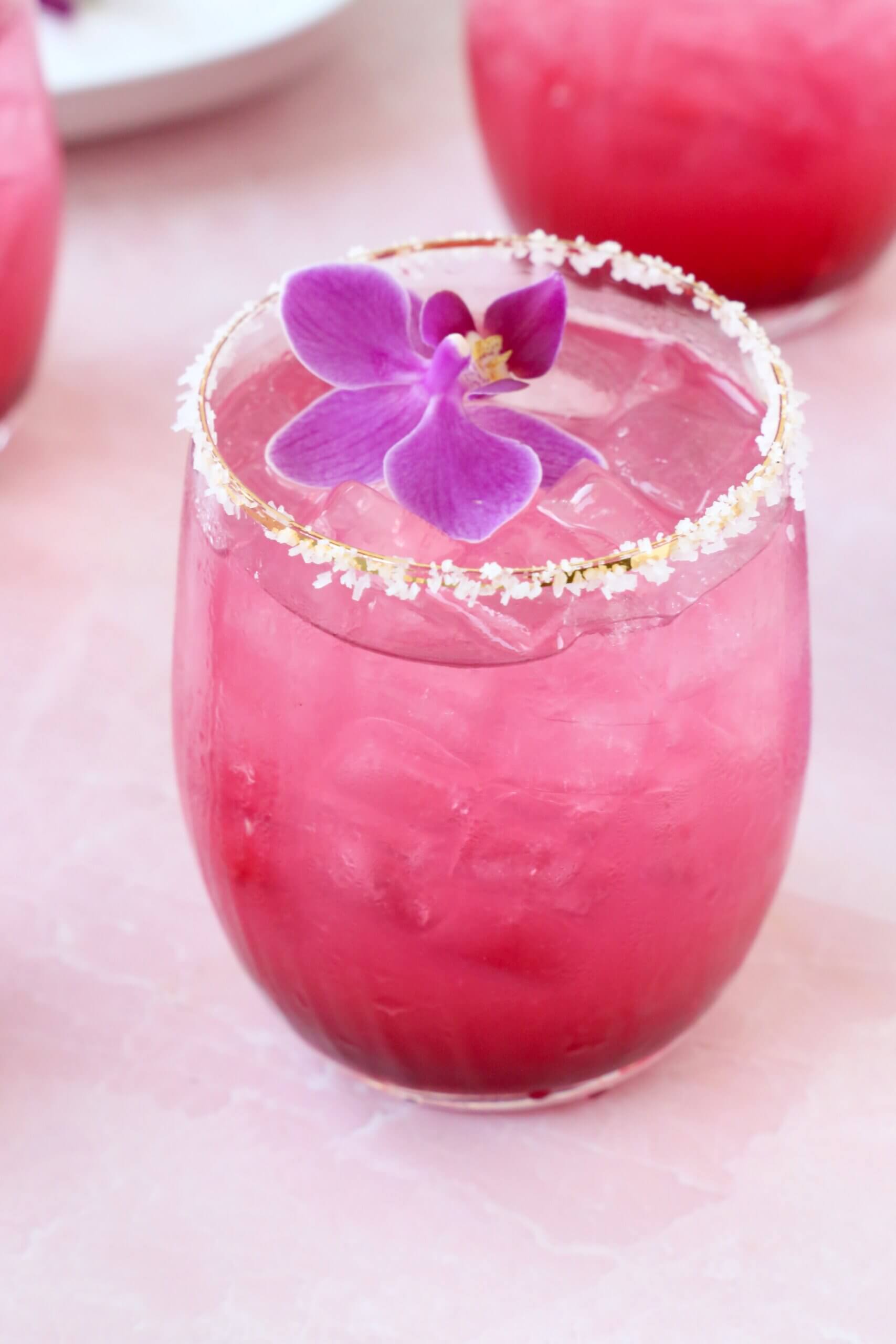 A clear glass with salt on the rim and a bright pink liquid and a purple orchid flower on top.