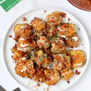 A white plate filled with light brown smashed potatoes topped with crumbled bacon, green chives and yellow cheese.