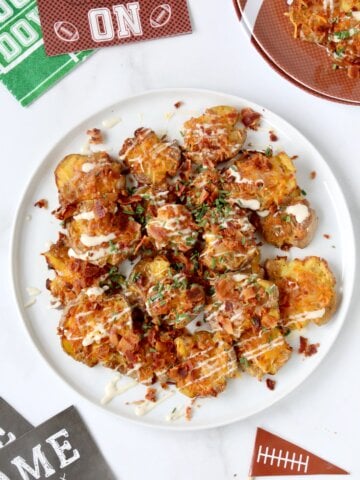 A white plate filled with light brown smashed potatoes topped with crumbled bacon, green chives and yellow cheese.