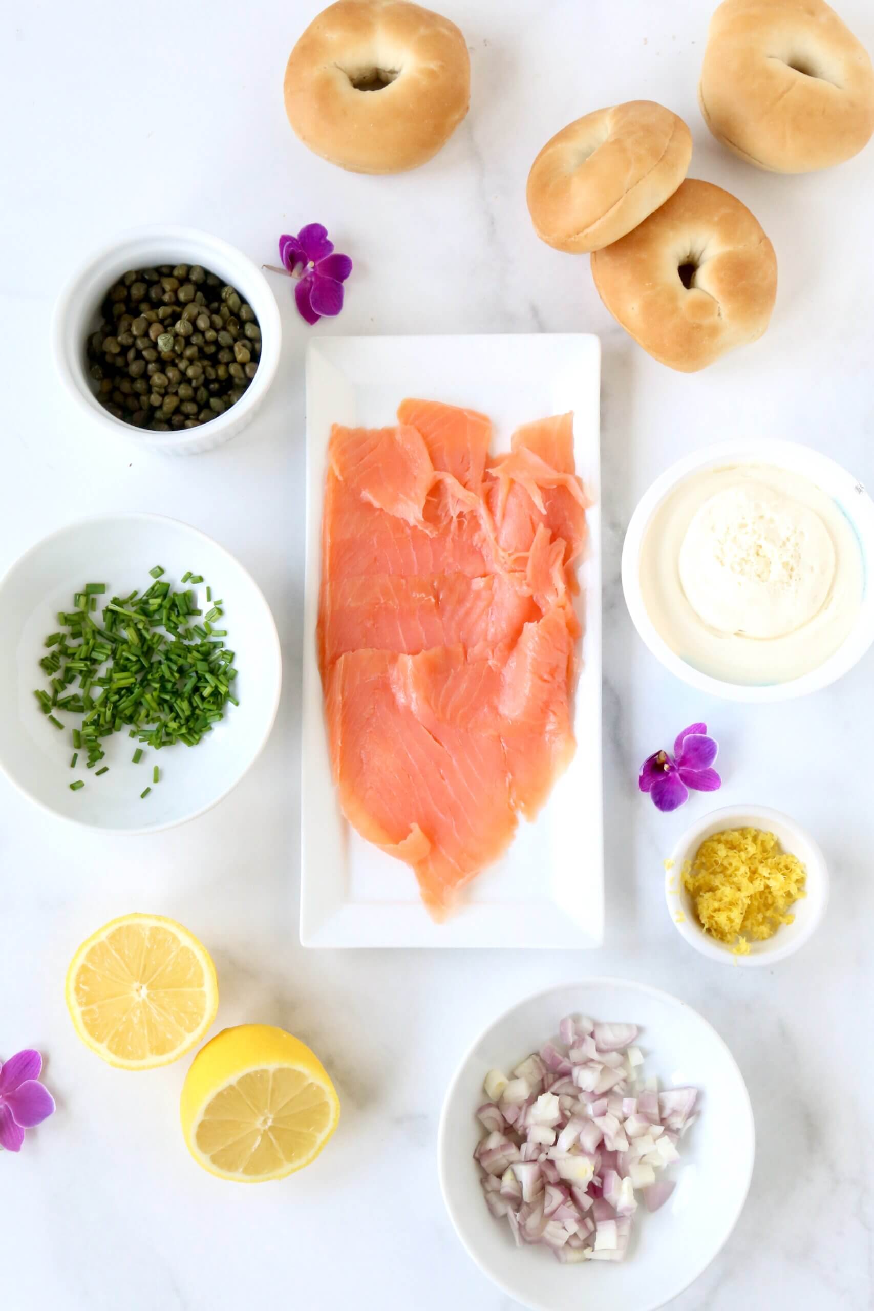 A white platter topped with orange colored smoked salmon surrounded by while bowls filled with green chives, yellow lemon zest, green capers and a lemon halved.