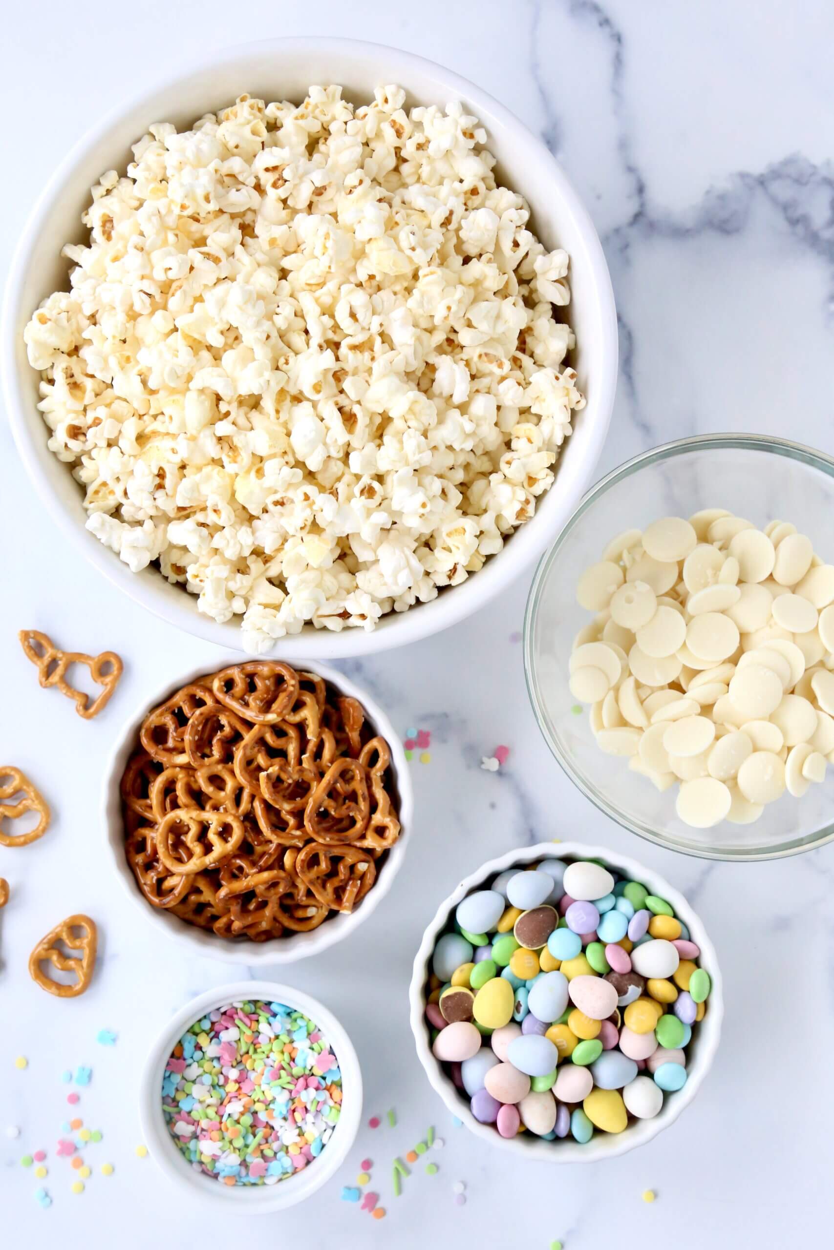 Five bowls filled with white popcorn, brown pretzels. white chocolate chips, pastel color m&m and colorful sprinkles.