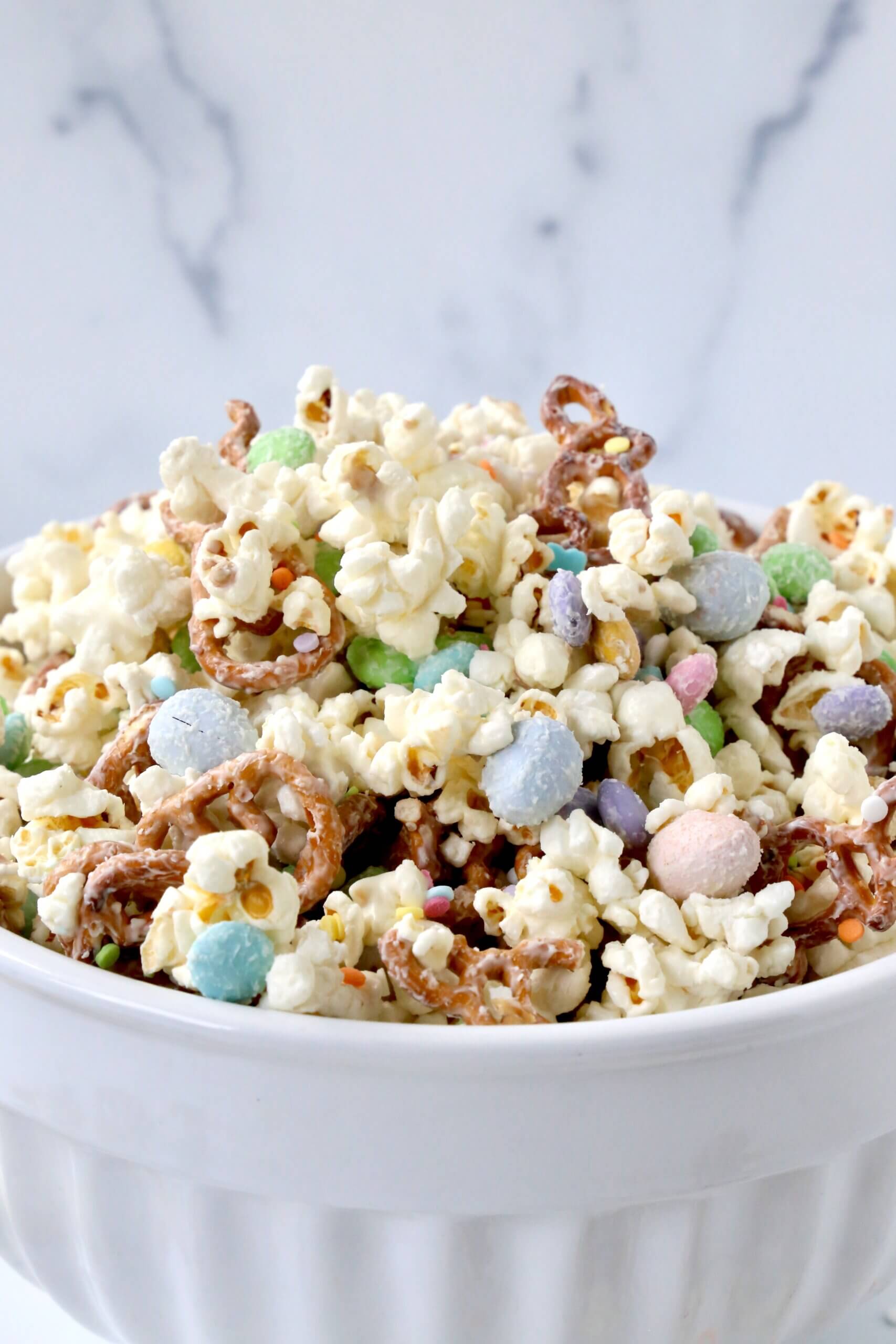 A white bowl with white popcorn, brown pretzels, pastel color m&m's.