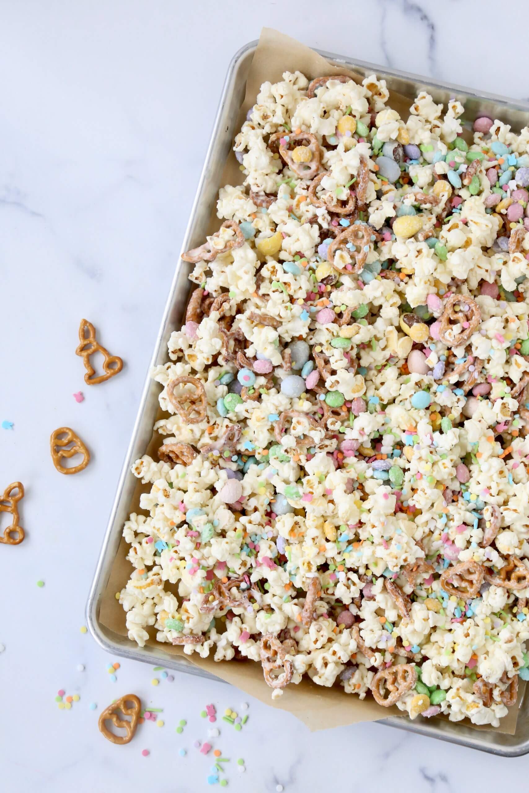 A silver tray with white popcorn, brown pretzels, colorful m&m's and colorful sprinkles.