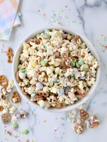 A white bowl with white popcorn, brown pretzels, pastel color m&m's.